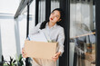 © kenchiro168 - Young Asian woman in white shirt holding cardboard box with office supplies, looking thoughtful and worried, standing outside modern office building after job loss