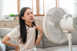 Suffer summer heat wave stroke, hot weather, tired asian young woman sitting on couch in living room without air-conditioner, using electric fan, wave fan to cool for ventilation when high temperature