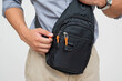 © Mdv Edwards - Close-up of a Southeast Asian man adjusting a black sling bag with orange zipper details, designed for urban or outdoor use. Isolated on a light background.