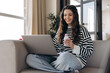 © Maria Vitkovska - Happy female student sitting on sofa using laptop and holding bottle with yogurt at home