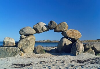 Aufkleber - A stone archway delicately balanced on a sandy beach, framing a tranquil ocean view under a vibrant blue sky.  The rocks vary in size and shape, creating a natural, artistic compositio : Generative AI