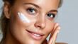 © mila103 - A young woman smiles gently as she applies moisturizer to her cheek, highlighting her radiant skin. Soft lighting enhances her natural beauty, creating a serene atmosphere