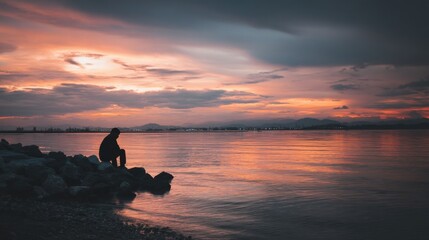 Poster - Silhouette of a person sitting on rocks by the sea during a vibrant sunset, contemplating the tranquil ocean and distant city lights. : Generative AI