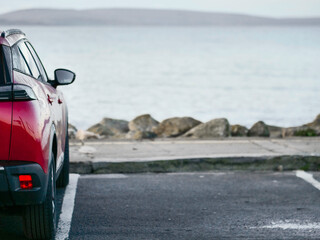 Wall Mural - Red car parked in car park with stunning view on nature scenery with ocean and mountains and cloudy sky. Silverstrand area, Galway city, Ireland. Rent motor for a tourist trip concept.