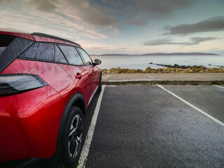 Wall Mural - Red car parked in car park with stunning view on nature scenery with ocean and mountains and cloudy sky. Silverstrand area, Galway city, Ireland. Rent motor for a tourist trip concept.
