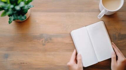 Poster - Overhead view of hands holding open blank notebook on wooden table, with a plant and coffee cup nearby, ready for writing or planning. : Generative AI