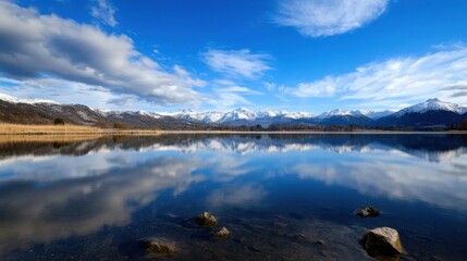 Aufkleber - Serene lake reflecting snow-capped mountains under a vibrant blue sky, with calm water and dark rocks in the foreground. : Generative AI