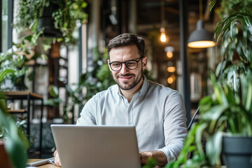 Poster - man is sitting at a table with a laptop in front of him. He is wearing glasses and smiling. The scene is set in a room with plants, giving it a relaxed and comfortable atmosphere