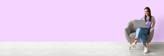 Poster - Positive young caucasian woman working online, sitting in armchair with coffee and using laptop against pink studio background, full length, panorama with copy space, mockup