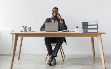 Poster - Cheerful black businessman in formal suit talking on smartphone, having conversation, sitting at table with laptop against white studio wall, copy space. Remote communication concept