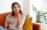 Pensive young woman holding smartphone and looking away while relaxing on couch at home, beautiful female deep in thoughts using mobile phone in domestic interior, thinking and planning something
