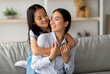 © Prostock-studio - Happy asian little girl hugging her beautiful mother and smiling to each other, sitting on sofa in living room, copy space. Adorable family young woman and her daughter embracing