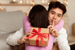 © Prostock-studio - Loving Asian Husband Hugging Wife Holding Christmas Gift Behind Her Back Preparing Surprise Celebrating Holiday At Home, Gesturing Hush Sign. Birthday Present And Holidays Celebration Concept