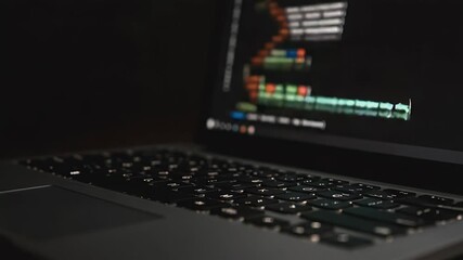 Close-up of a laptop keyboard illuminated in a dark room, showcasing coding activity on the screen in the background