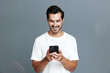 © SHOTPRIME STUDIO - Smiling young man wearing a white t-shirt, looking at his mobile phone with a joyful expression against a plain gray background. People technology lifestyle concept.