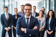 © soem - A smiling man in a suit standing in front of a group of business professionals in a modern office setting.
