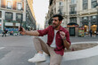 © BullRun - cheerful male traveler sits in the middle of a city street, laughing and gesturing while talking on video call with smartphone. lively urban background adds to the energetic atmosphere of his journey