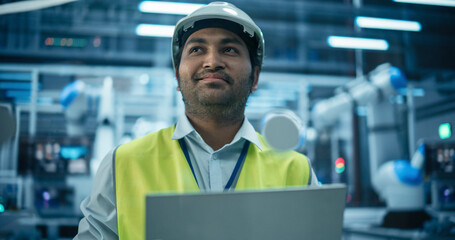 Fototapete - Indian Engineer Working in a High Tech Factory with Robotic Arms Assembling Electronic Components on a Production Line Conveyor Belt. South Asian Man Wearing a High Visibility Vest and Using a Laptop