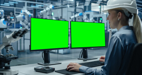 Fototapete - Female Administrator Working Behind a Desk in a Modern Industrial Facility with Automated Robotic Arms. Industrial Specialist Working on a Computer with Green Screen Mock Up Template