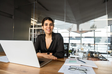 Portrait young it specialist latin hispanic business lady working online on laptop pc sitting at desk in modern office smiling at camera. Middle eastern indian woman using computer at work. Copy space