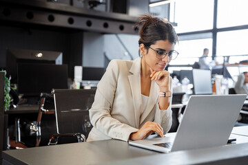 Young professional specialist latin hispanic business lady working on laptop pc sitting at desk in modern office space. Busy middle eastern indian woman using computer technology app for work online
