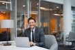 © ngalong project - Smiling man in suit sits at desk with laptop in office environment.