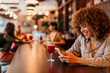 © La Famiglia - Cheerful young woman using smartphone and drinking juice at bar counter