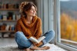 © Garik - Young woman enjoys reading a book in cozy sweater while sitting by large window with autumn view