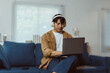 © Crystal - Young man using laptop and headphones while working remotely from home, sitting comfortably on couch