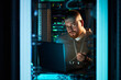 © DragonImages - Man working in server room, analyzing data on laptop surrounded by racks of servers, maintaining focused expression throughout work setting part of technology maintenance