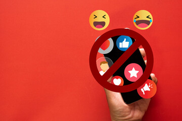 Concept of no social media interaction,Social Media Detox,Social Detoxification.,Hand holding a smartphone with crossed-out among  social media notifications and emoji icons on red background.