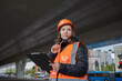 © night87 - A female civil engineer in an orange construction helmet and vest stands under an overpass and holds a tablet in her hands.