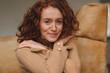 © Iryna - Young woman with curly red hair smiles warmly while sitting comfortably on couch in cozy indoor setting during afternoon
