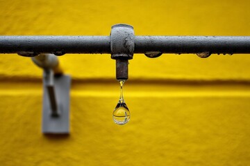 Poster - Water droplet forms and drips from a metallic pipe against a vibrant yellow wall