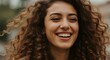 © Image - Close-up portrait of a smiling woman with curly brown hair, radiating warmth and joy, captured in a natural outdoor setting.