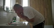 © Marco - Man rinsing face under running water, leaning forward at sink, hands cupped to splash water, bathroom environment with wooden elements, morning grooming ritual