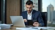 © Nathan - A focused Caucasian man in a suit works diligently on a tablet, while sitting at his desk with a laptop in a modern office.