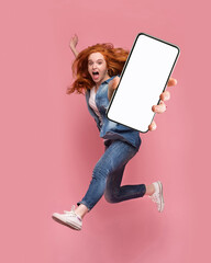 Poster - Excited redhead woman having fun, jumping,demonstrating mobile phone with empty white screen on pink studio background. Creative mockup collage with space for website or app design