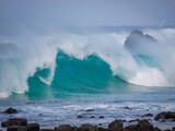 Wave of summer swell crashes with immense force against volcanic rocks on coastline as misty offshore spray rises into the air, showcasing raw power of the sea and untamed nature of Atlantic Ocean.