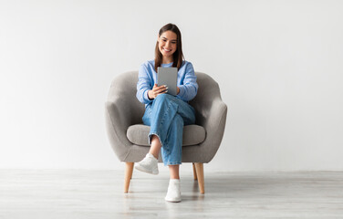 Poster - People, technology and communication concept. Portrait of smiling young woman with digital tablet sitting in armchair, working online against white studio wall, full length