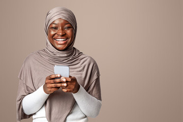 Poster - Joyful Black Muslim Lady In Hijab Holding Smartphone And Laughing At Camera, Enjoying Mobile Communication And Online Shopping, Browsing New App, Standing On Light Studio Background, Copy Space