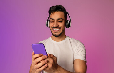 Poster - Portrait of cool young Arab man in wireless headphones using smartphone in neon light. Millennial middle Eastern guy checking new mobile app, listening to music, watching video, chatting online