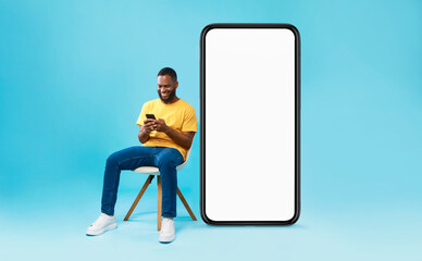 Poster - Cheerful young black man sitting on chair near big smartphone with empty screen, using mobile device, presenting space for website, advertisement or mobile app, blue studio background