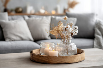 Poster - Cozy living room scene featuring a wooden tray with lit candles, a glass vase of dried cotton flowers and pampas grass, and a blurred grey sofa in the background. : Generative AI