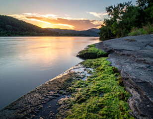 Wall Mural - serene river landscape at sunset, featuring rocky shore covered in vibrant green algae. calm water reflects warm hues of sky, creating peaceful atmosphere