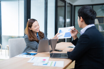 Poster - Two asian businesspeople are discussing and analyzing financial charts and data using laptops and documents, collaborating on a project in a modern office environment