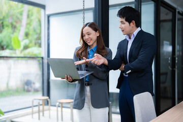 Poster - Two Asian business professionals standing together in a modern office, discussing work while using a laptop, collaborating on a project and exchanging innovative ideas in a bright, elegant space