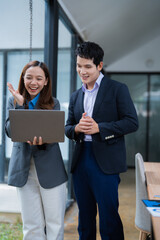 Fototapete - Two asian business people wearing suits are standing in the office, looking at a laptop and having a video conference, smiling and gesturing