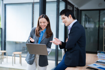Aufkleber - Two Asian businesspeople smiling and engaging in discussion while working on a laptop in a modern office setting, exemplifying collaboration and teamwork in a corporate environment