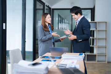 Poster - Two Asian business professionals standing together in a modern office, discussing a document and exemplifying teamwork and collaboration in a corporate environment